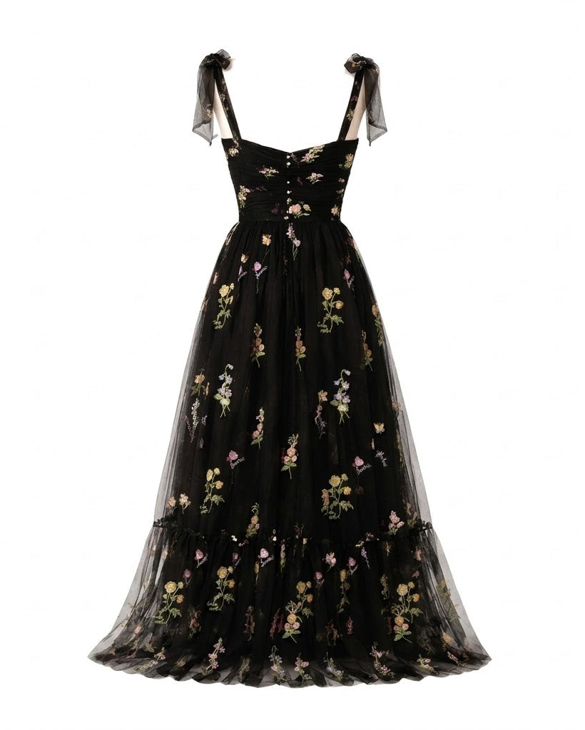 A black midi-length A-line dress made of sheer tulle, decorated with a delicate multicolor floral print, featuring a sweetheart neckline and sheer shoulder straps tied at the shoulders.
