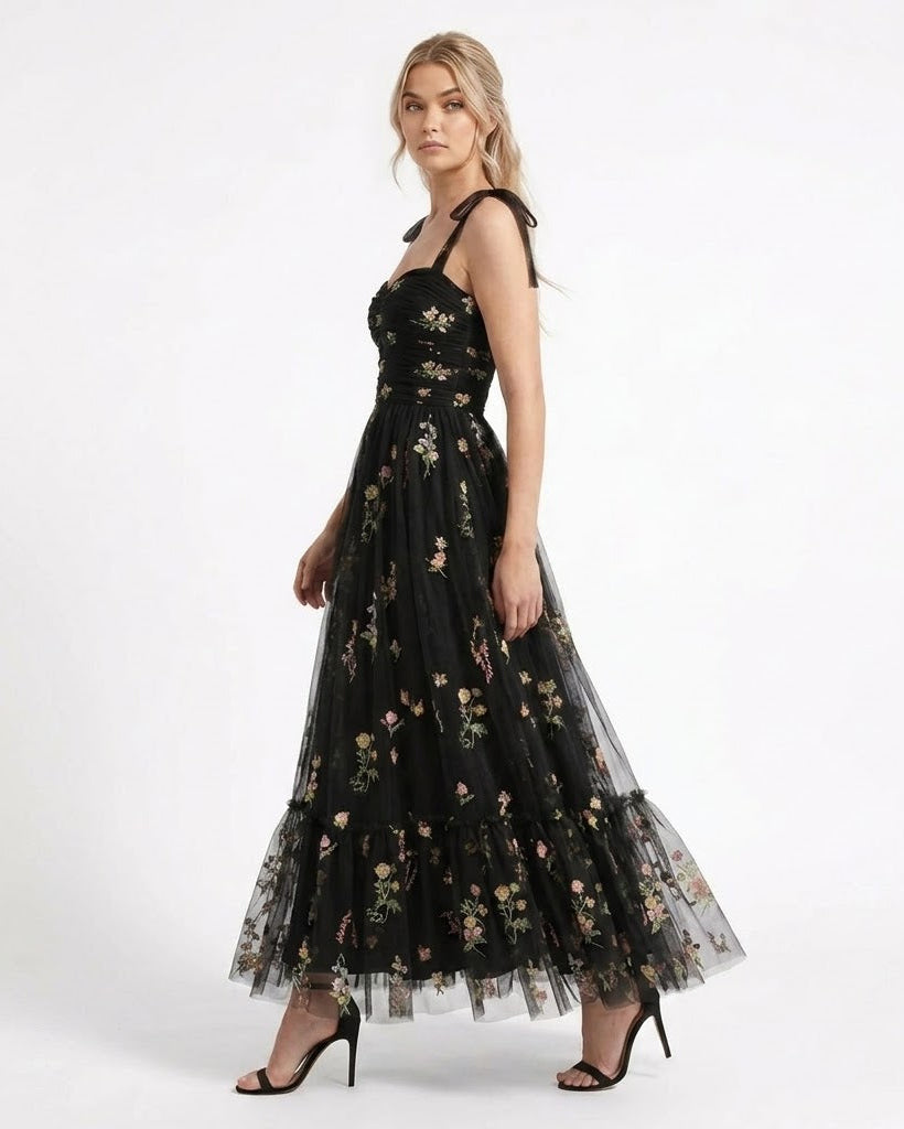 A woman modeling a black sleeveless maxi dress with floral embroidery and sheer tulle layers, paired with black high-heeled sandals, against a plain white studio background.