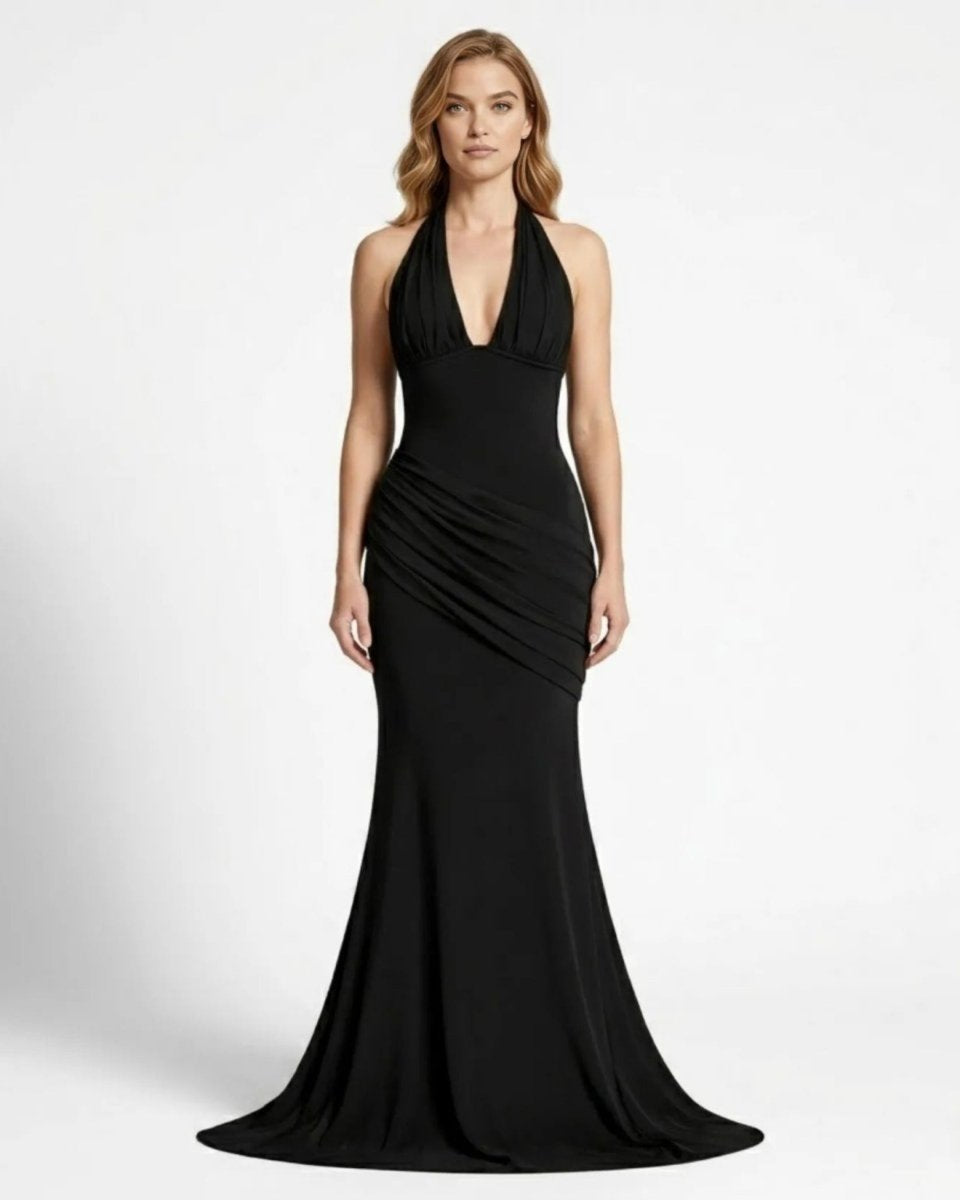 A woman modeling a floor-length black halter gown with a deep V-neckline and draped ruching at the waist, photographed against a plain light gray studio background.