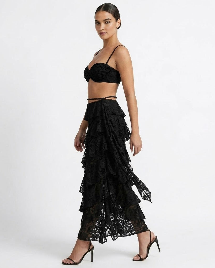 A woman modeling a black lace two-piece outfit consisting of a bandeau-style crop top and a long, tiered ruffled maxi skirt, paired with black strappy high-heeled sandals against a plain white studio background.