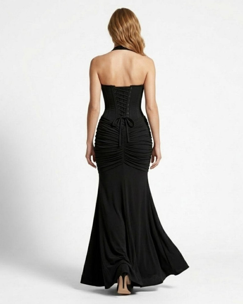 A woman viewed from the back wearing a strapless black formal gown with a corseted waist, gathered ruching, and a flared skirt, photographed against a plain light gray studio background.