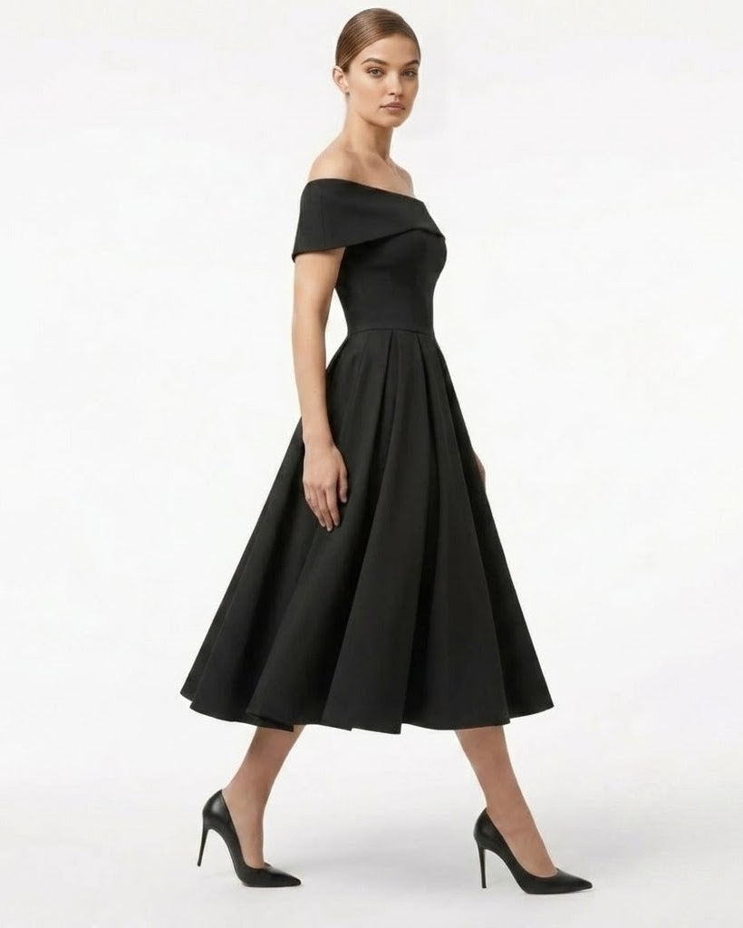 A woman modeling a black off-the-shoulder midi dress with a flared skirt and black pointed-toe heels against a plain white studio background.