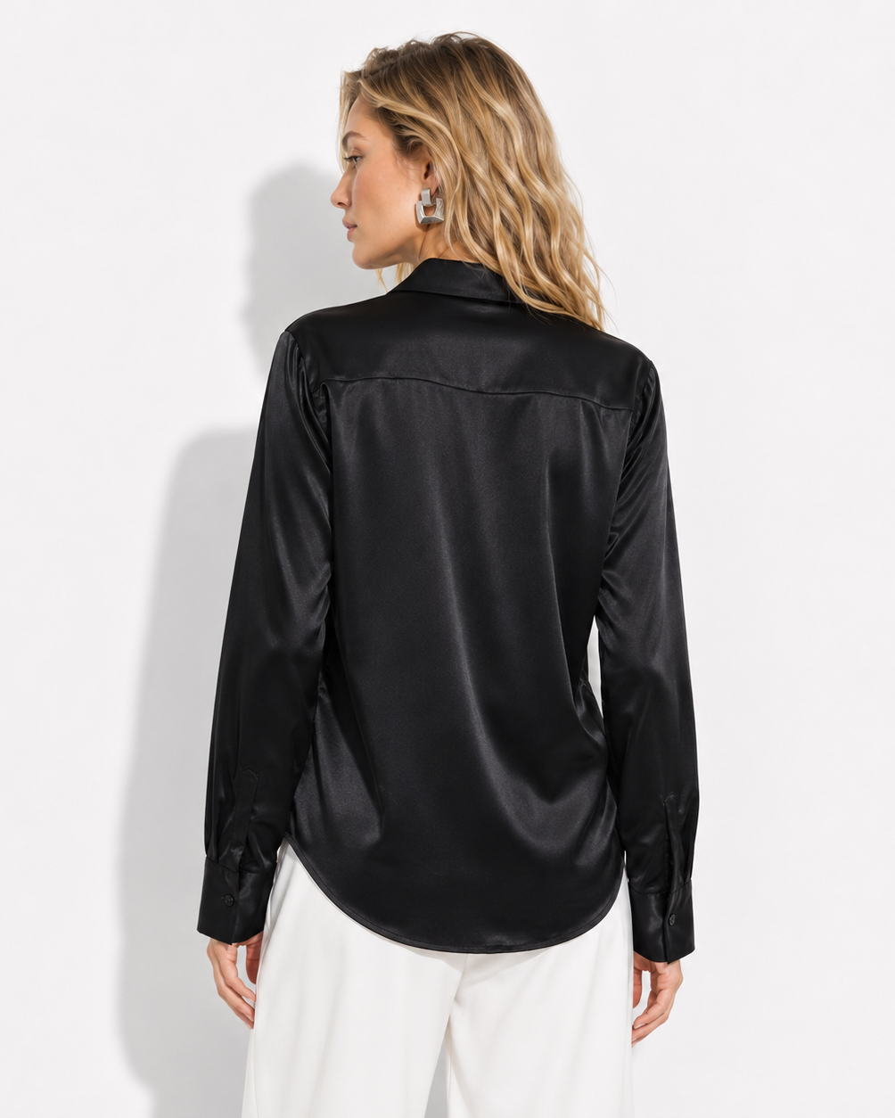 A woman shown from the back wearing a black satin long-sleeve button-up shirt and white trousers, standing against a plain white studio background.
