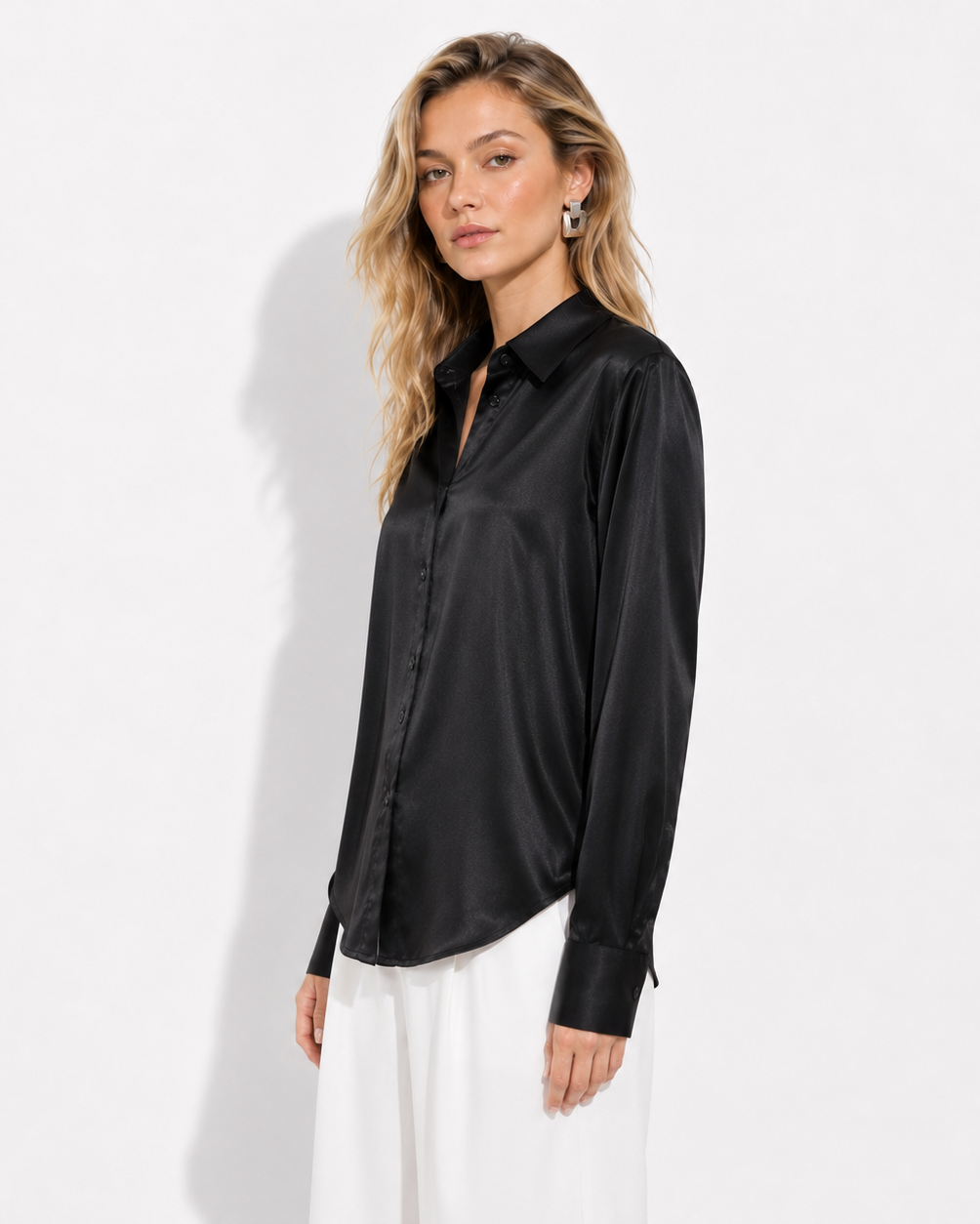 A woman modeling a black satin long-sleeve button-up shirt with white trousers, standing against a plain white studio background.