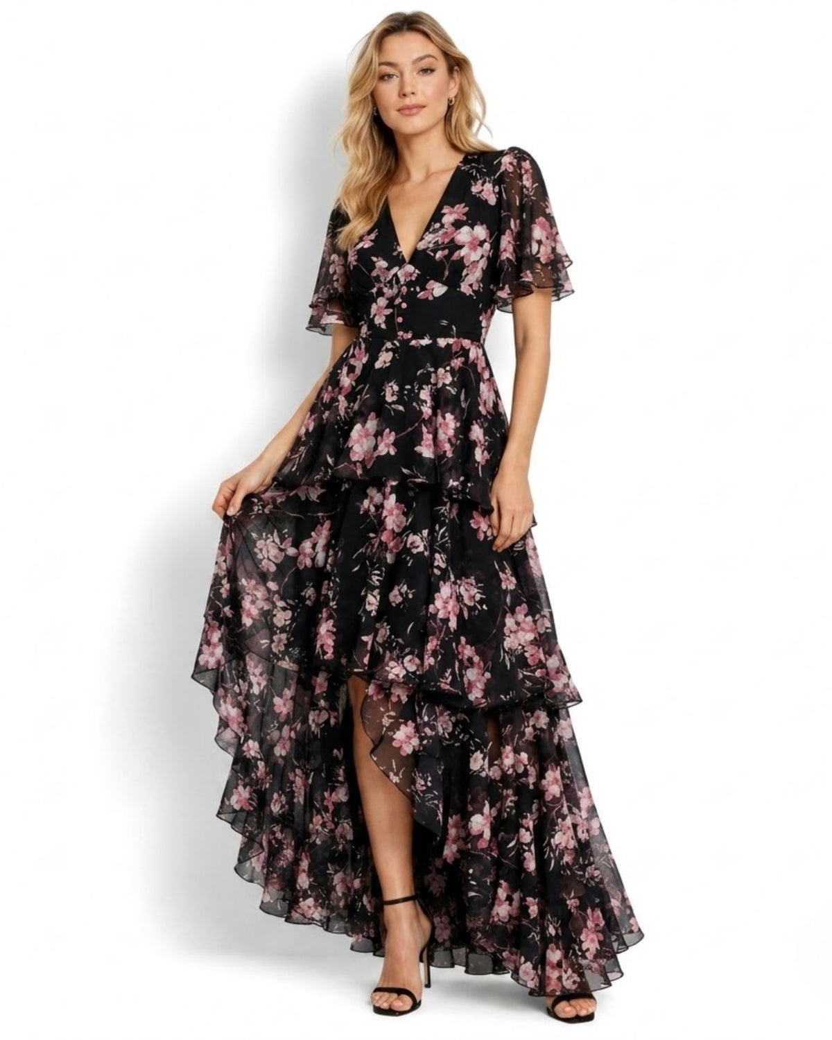 A woman modeling a black high-low midi dress with a pink floral print, ruffled short sleeves, and a tiered skirt, set against a plain white background.