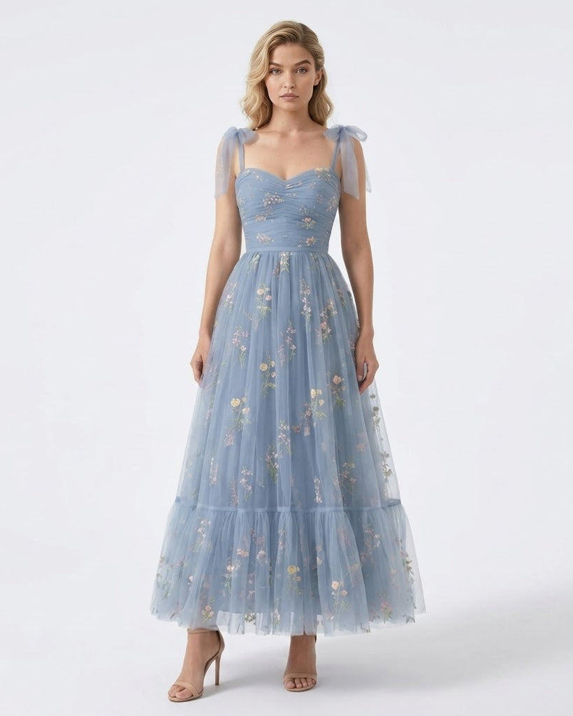 A woman modeling a light blue tulle maxi dress with floral embroidery, a sweetheart neckline, and sheer shoulder straps, photographed against a plain white studio background.