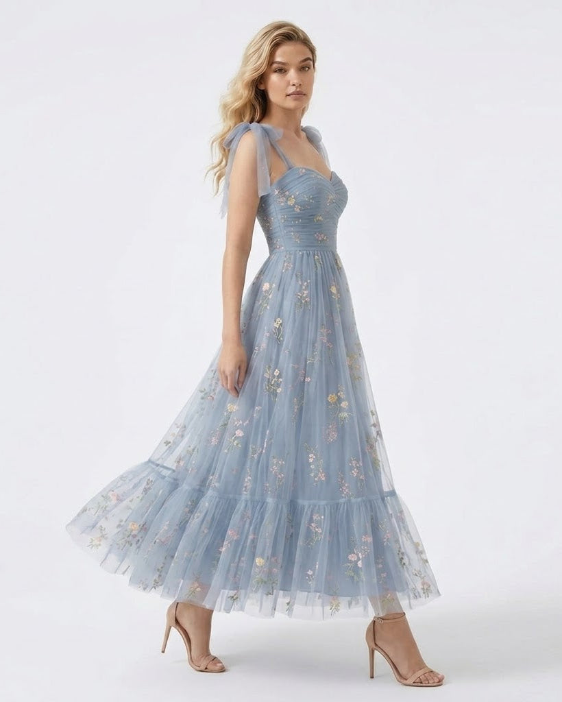 A woman modeling a light blue sleeveless maxi dress with a fitted bodice, thin shoulder straps, and a tiered tulle skirt adorned with small floral embroidery, posed against a plain white studio background.