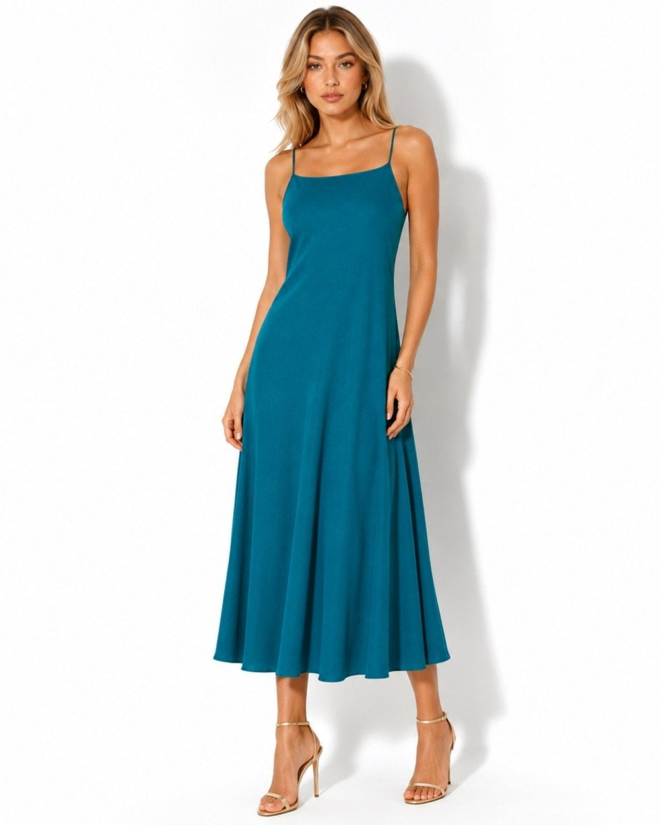 A woman modeling a teal blue midi-length spaghetti strap dress with a flared skirt, paired with nude high-heeled sandals, against a white studio background.
