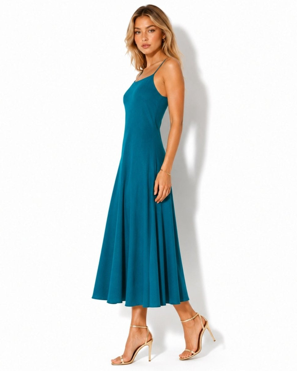 A woman modeling a sleeveless teal maxi dress with a fitted bodice and flared skirt, paired with clear-strap high-heeled sandals, against a plain white studio background.