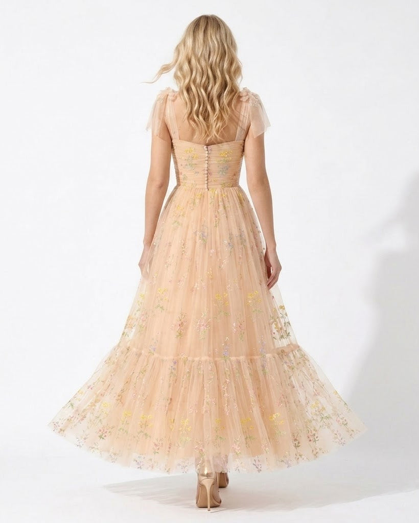 A woman viewed from behind wearing a sleeveless, tiered tulle maxi dress in a pale peach-beige color with delicate yellow and pink floral embroidery, set against a plain white studio background.