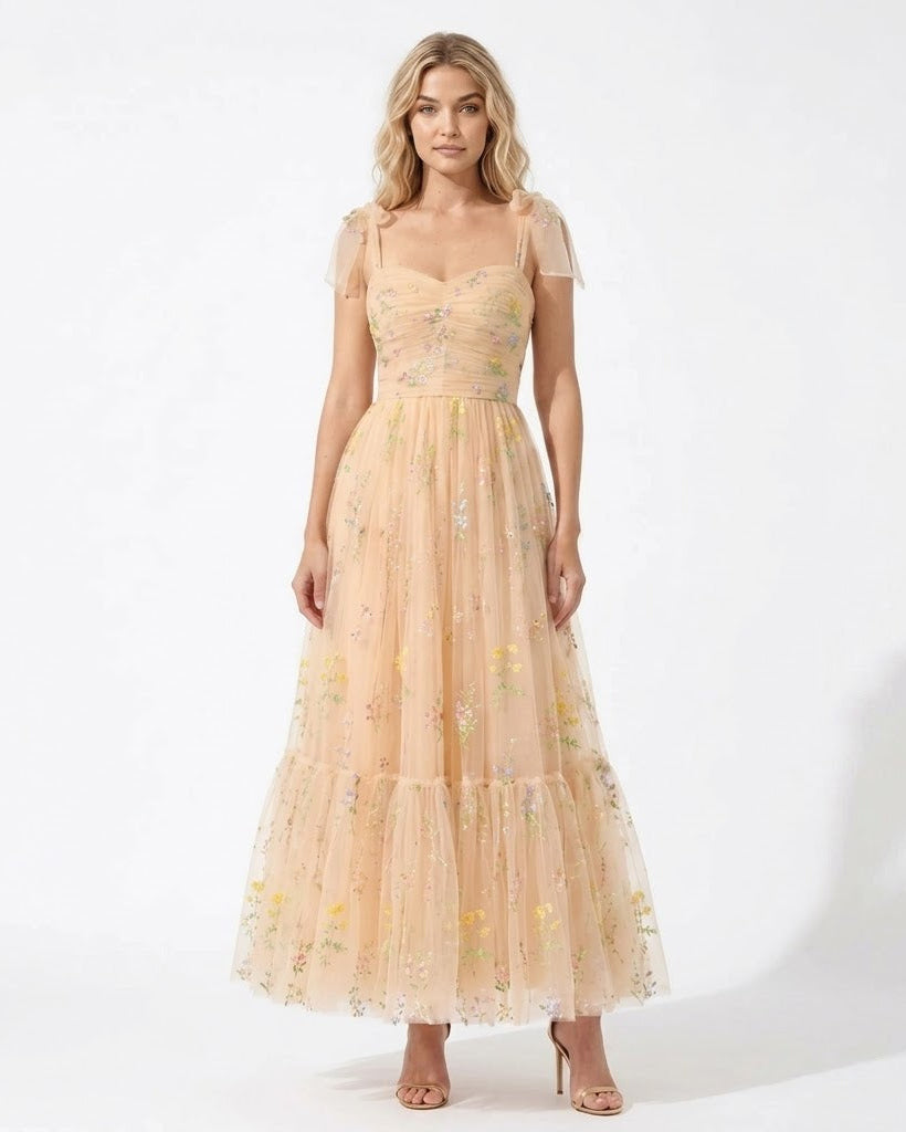A woman modeling a peach-beige tulle maxi dress with floral embroidery and sheer cap sleeves against a white studio background.