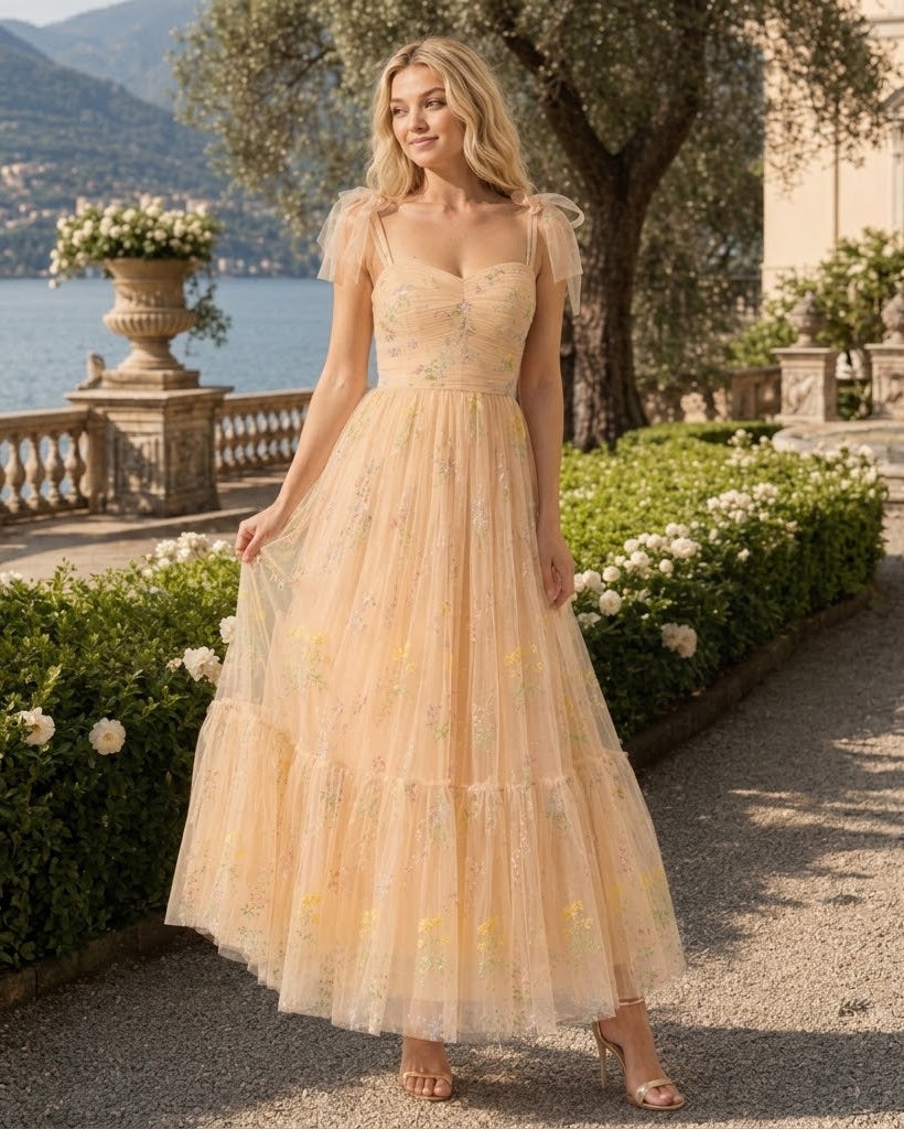 A woman stands outdoors on a stone path beside a lake, wearing a flowing pale peach maxi dress with sheer flutter sleeves, a fitted bodice, and a tiered tulle skirt with embroidered floral details.