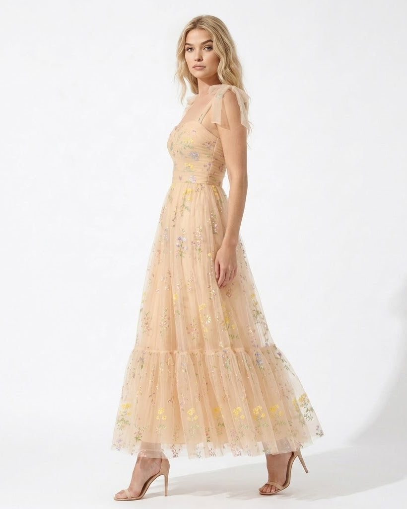 A woman models a sleeveless, tiered maxi dress in a light peach-beige tulle with delicate multicolor floral embroidery, paired with nude high heels against a white studio background.