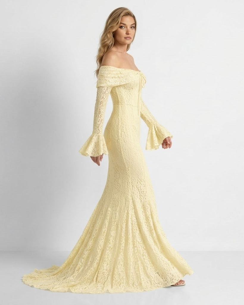 A woman modeling a pale yellow off-the-shoulder lace maxi gown with long flared sleeves and a fitted mermaid silhouette, photographed against a plain light gray studio background.