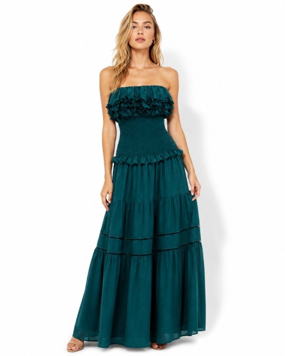 A woman modeling a strapless dark teal green maxi dress with ruffled details on the bodice and a tiered skirt, photographed against a plain white studio background.