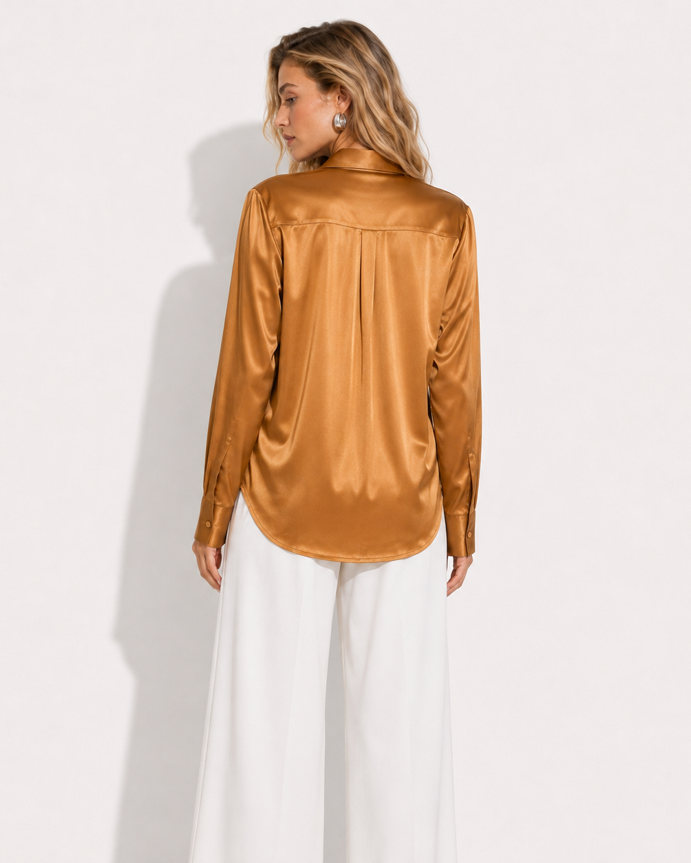 A woman shown from the back wearing a shiny bronze-brown satin long-sleeve blouse tucked into white wide-leg pants, standing against a plain white wall.