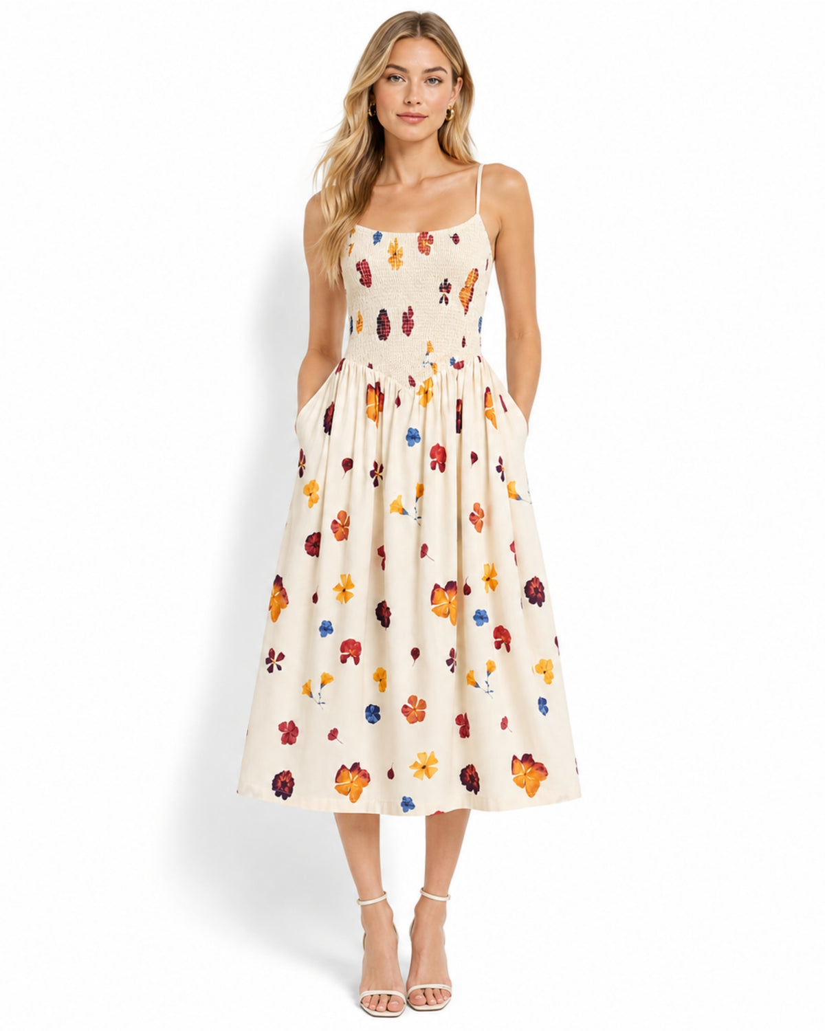 A woman modeling a sleeveless midi dress with thin straps, side pockets, and an allover multicolor floral print on a cream background, photographed against a plain white studio background.