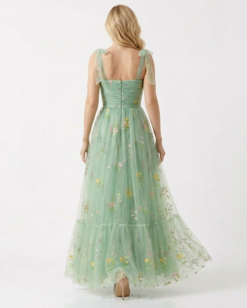 A woman viewed from the back wearing a long mint green tulle dress with floral embroidery and sheer shoulder straps, photographed against a plain white studio background.