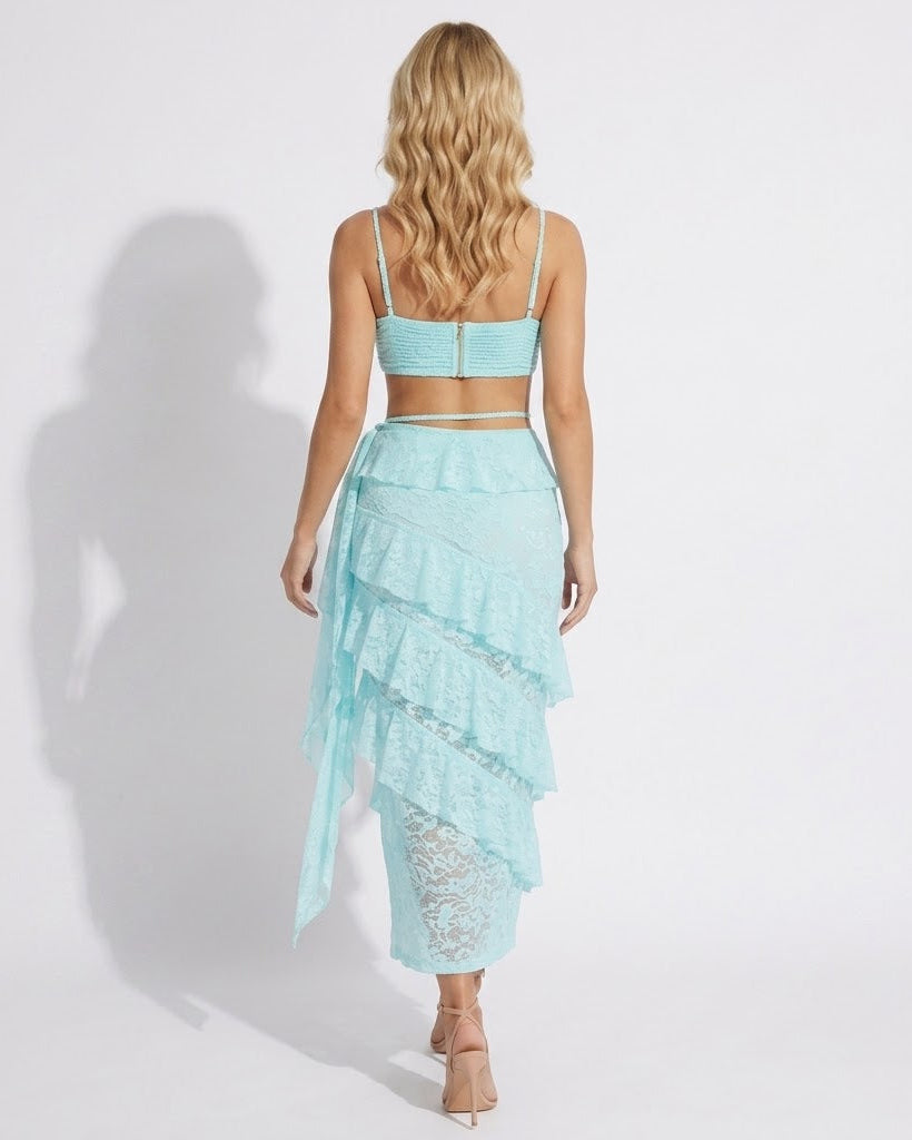 A woman shown from the back wearing a light aqua two-piece set consisting of a spaghetti-strap crop top and a matching tiered ruffled skirt with lace inserts, set against a plain white studio background.