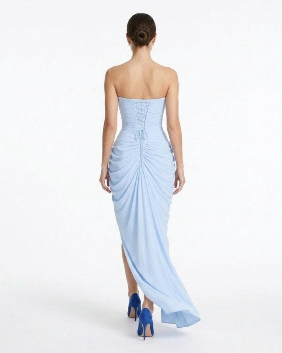 A woman shown from the back wearing a strapless light blue floor-length gown with ruching and a side slit, paired with blue heels, against a plain white studio background.