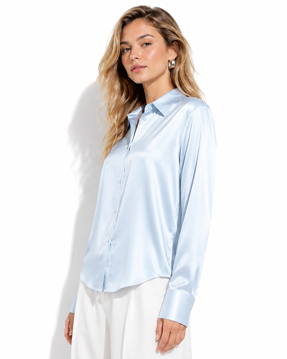 A woman modeling a light blue satin long-sleeve button-up shirt with white pants, photographed against a clean white studio background.