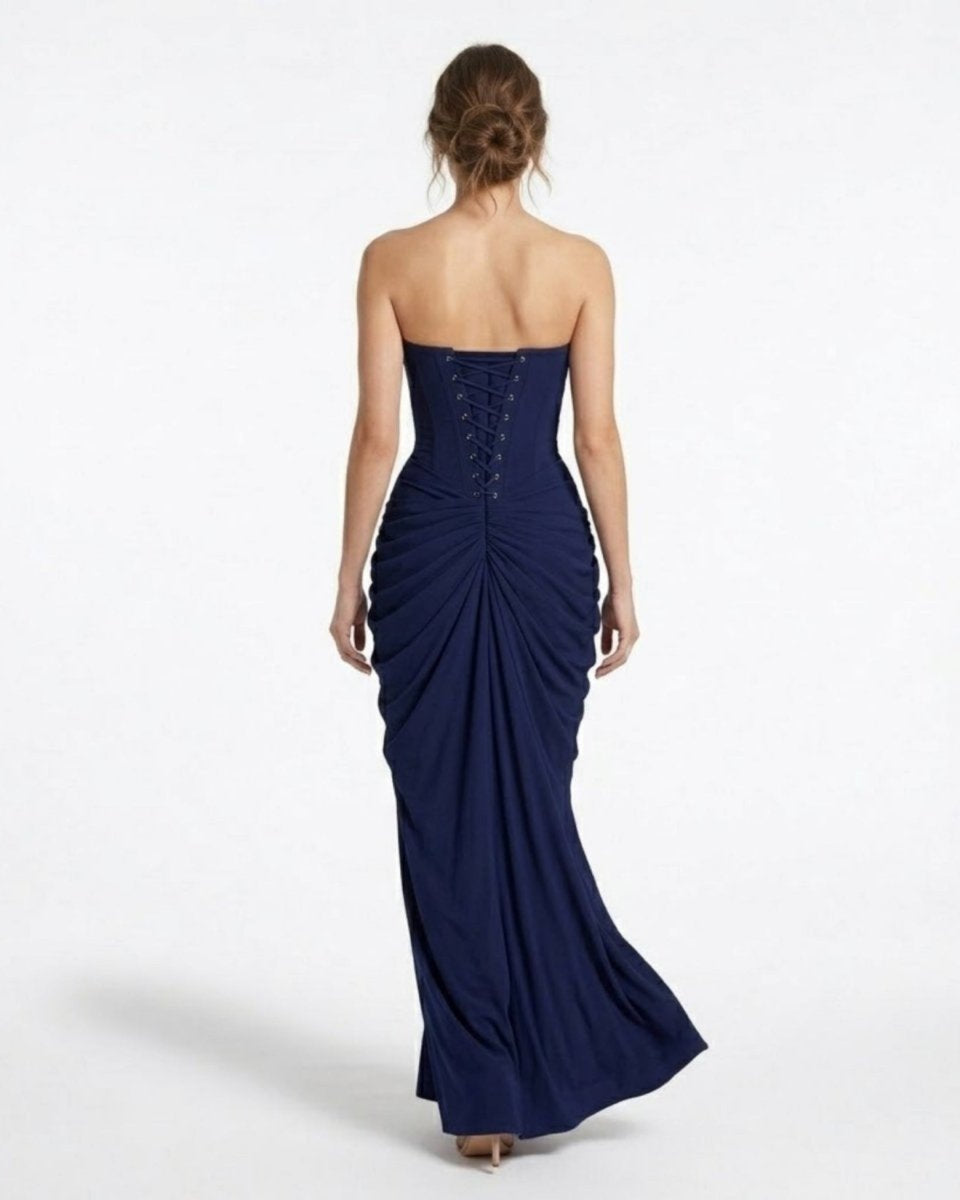 A woman viewed from the back wearing a strapless navy blue gown with a lace-up corset-style back and gathered skirt, photographed against a clean white studio background.