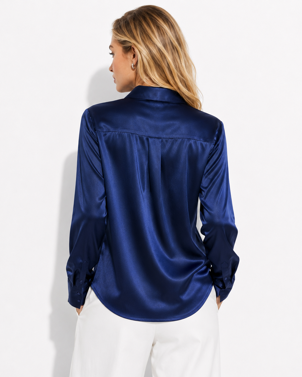 A woman viewed from the back wearing a shiny navy blue satin button-up shirt and white pants, standing against a plain white studio background.