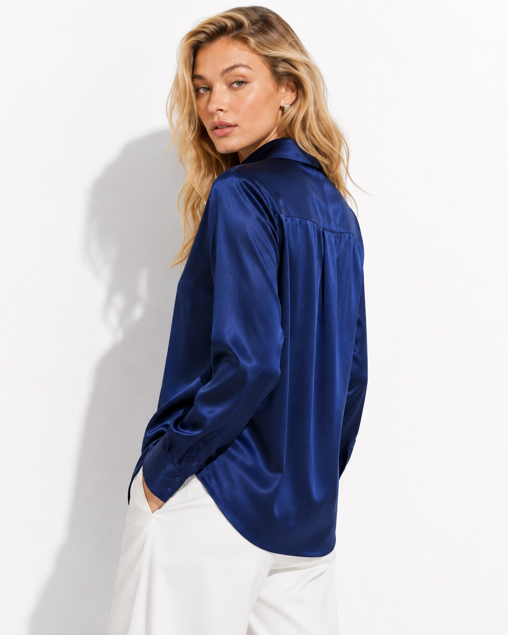 A woman wearing a shiny royal blue satin button-up shirt and white trousers, photographed against a clean white background with a soft shadow.