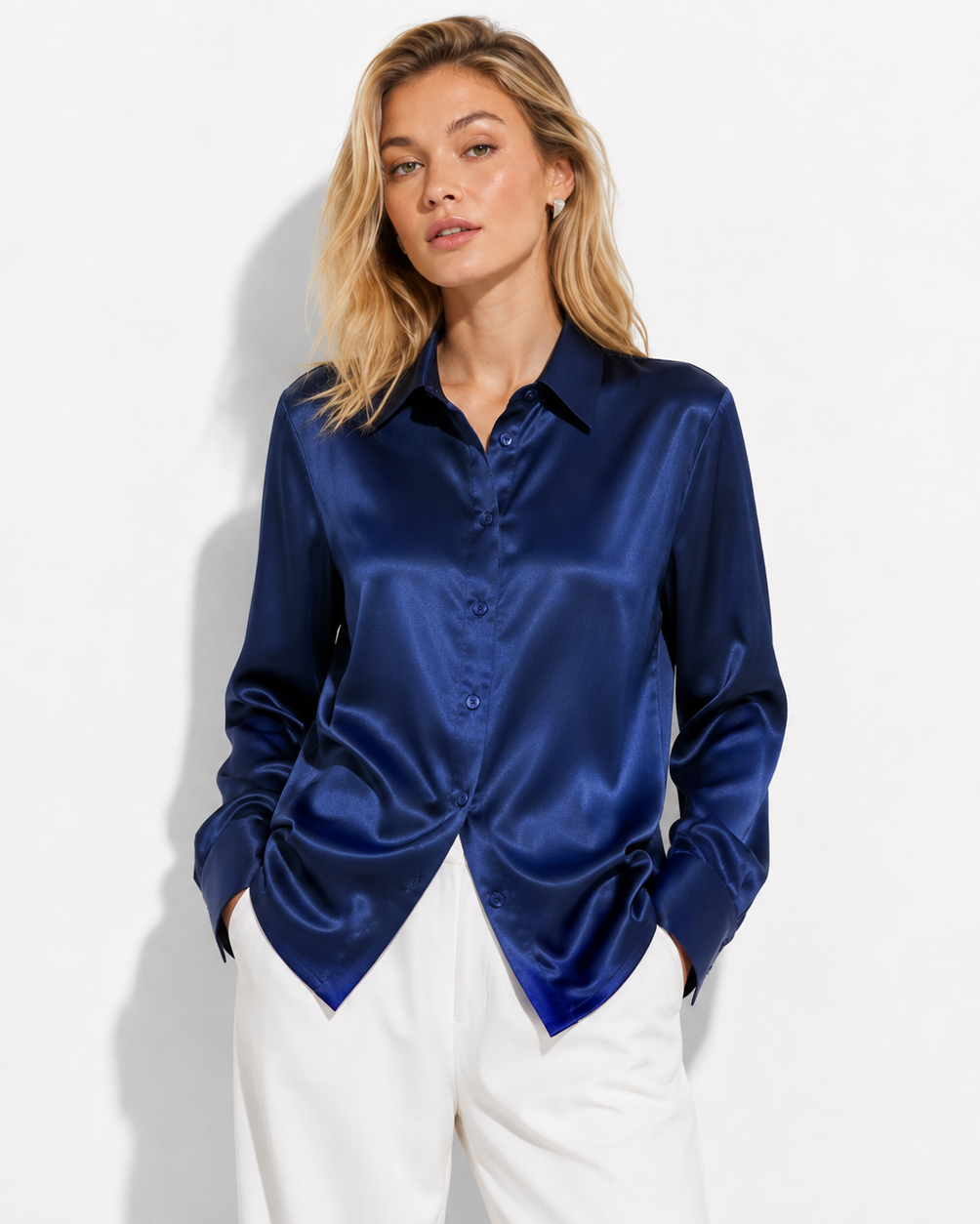 A woman modeling a long-sleeve satin button-up blouse in deep navy blue, paired with white trousers, against a plain white studio background.