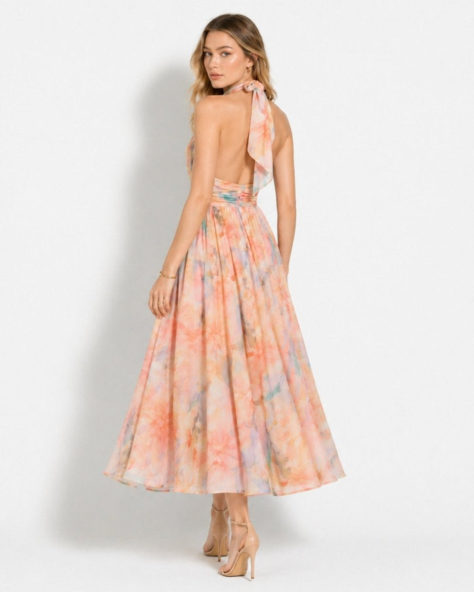 A woman modeling a one-shoulder maxi dress with a colorful tie-dye pattern in shades of pink, orange, and blue, shown from a back-three-quarter angle against a light gray studio background.