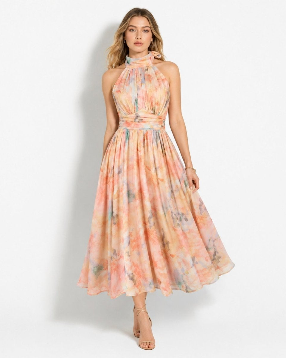 A woman modeling a sleeveless halter-neck midi dress with a flowing skirt, featuring a pastel watercolor tie-dye pattern in shades of peach, orange, pink, and light blue on a white studio background.