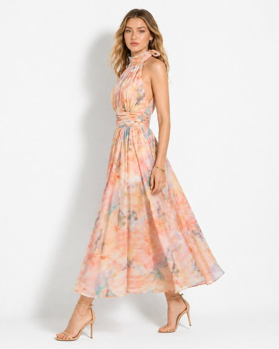 A woman modeling a sleeveless halter-neck maxi dress with a pastel tie-dye pattern in pink, orange, and yellow, paired with nude high-heeled sandals against a white studio background.