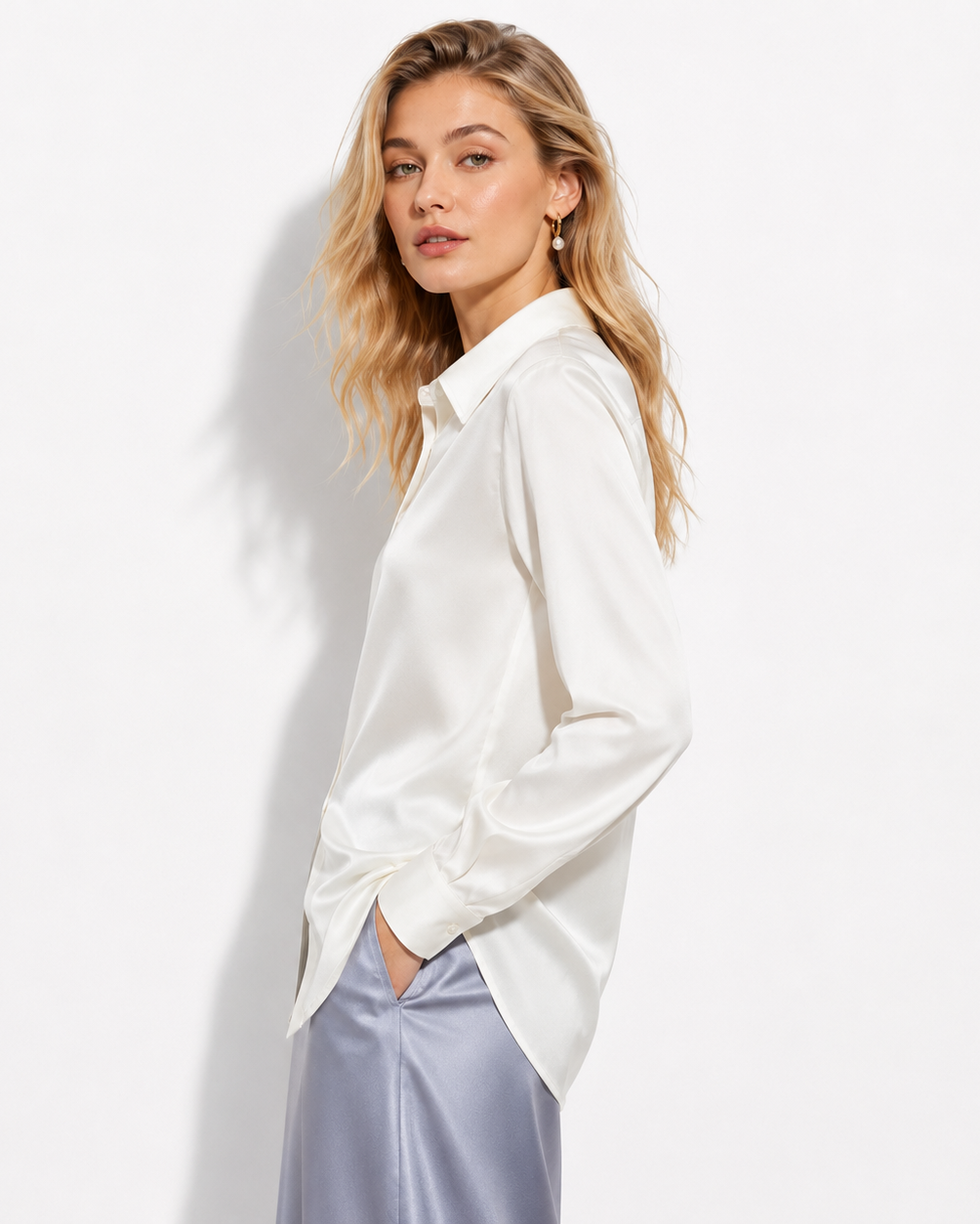 A woman wearing a white satin long-sleeve button-up shirt and light blue satin trousers, posing against a white studio background.