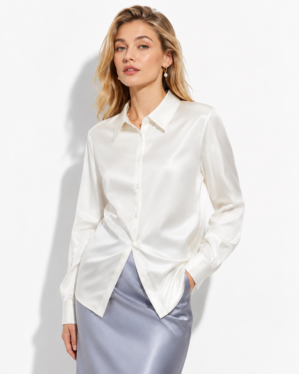 A woman modeling a white satin button-down shirt with a pointed collar and long sleeves, paired with a shiny light gray skirt, against a clean white studio background.