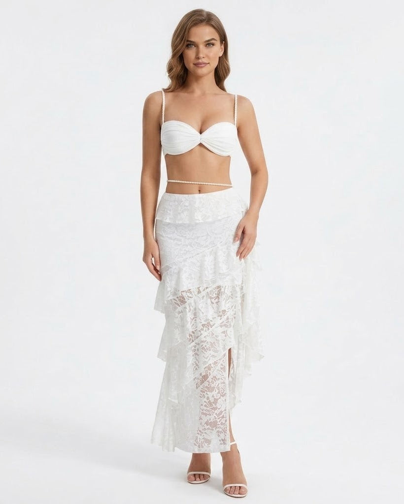 A woman modeling a white two-piece swimwear set featuring a textured bandeau-style bikini top and a ruffled lace maxi skirt, set against a plain white studio background.