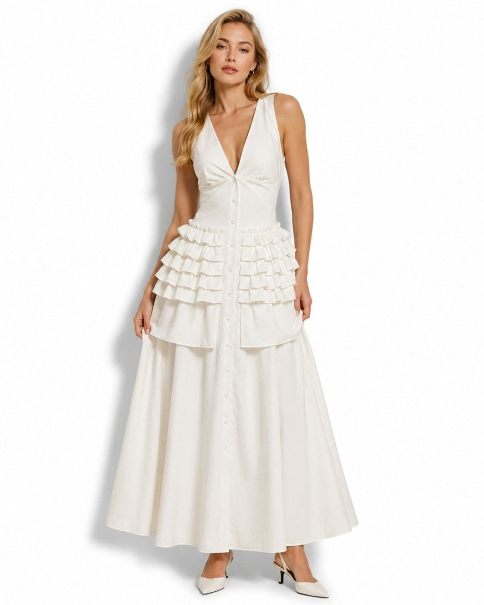 A blonde model wearing a sleeveless white maxi dress with a deep V-neckline, button-front bodice, tiered ruffle waist, and full skirt, paired with white pointed heels against a plain white studio background.