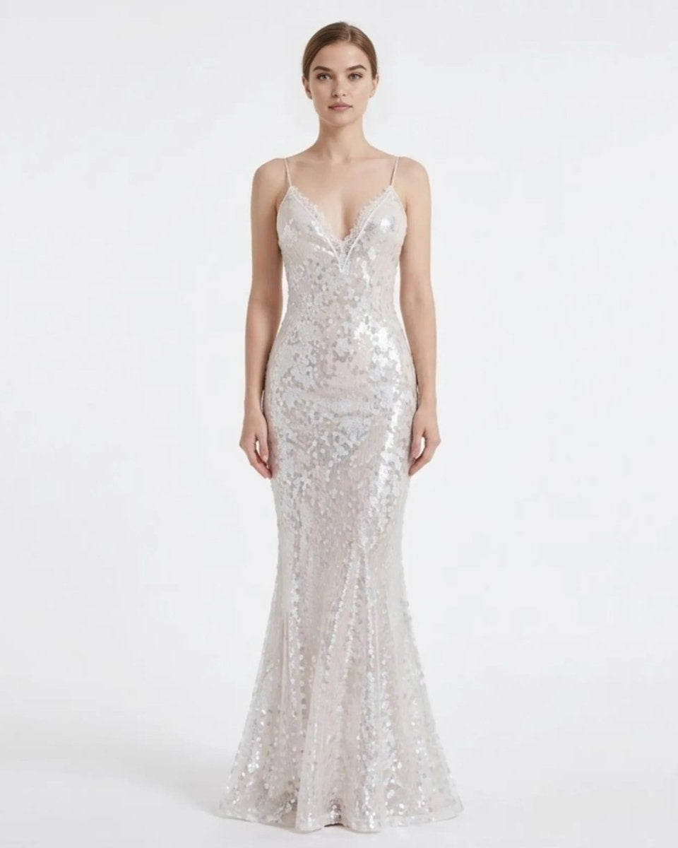A woman modeling a floor-length, spaghetti-strap mermaid gown covered in shimmering silver sequins against a plain light gray studio background.
