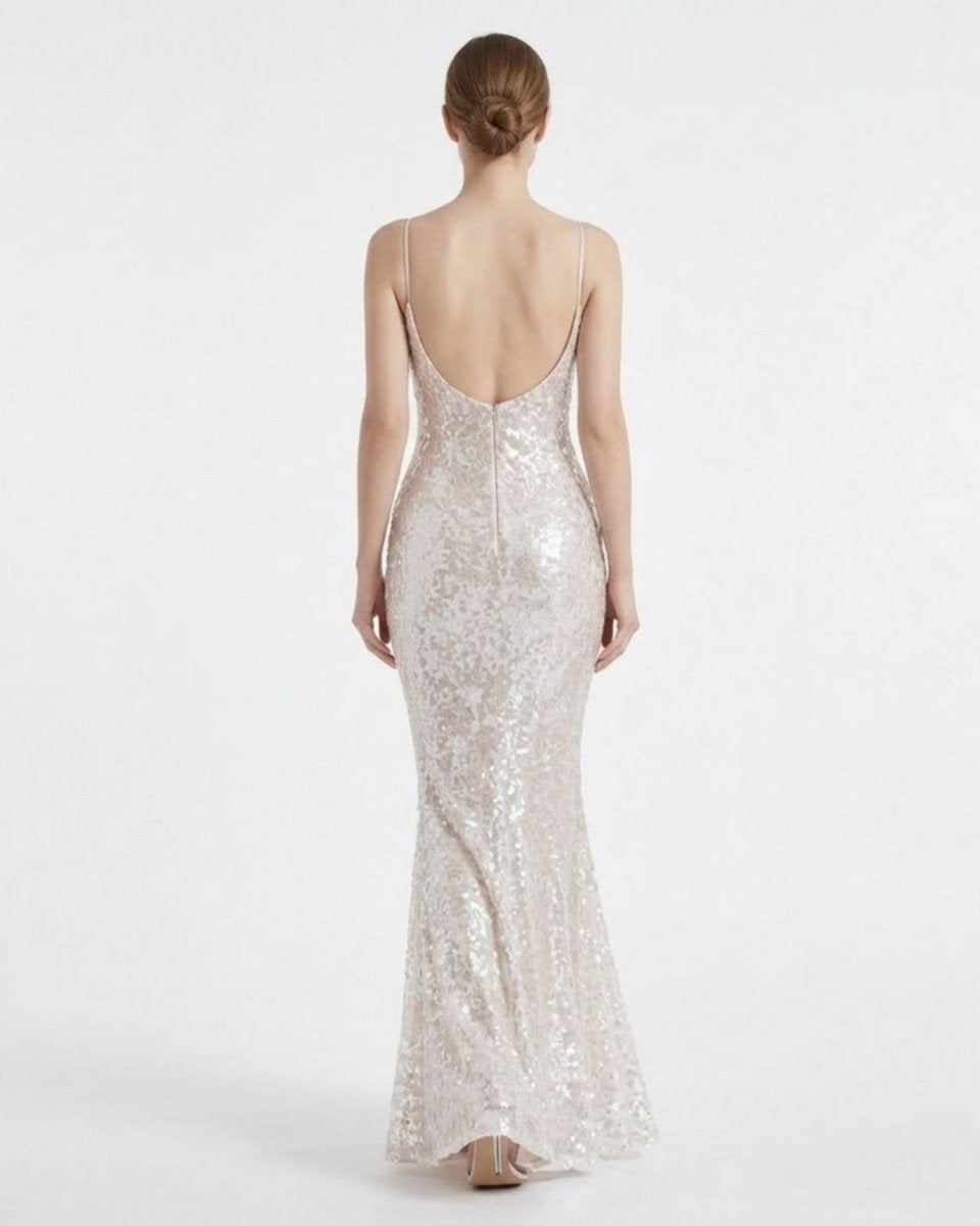 A woman viewed from the back wearing a floor-length, sleeveless evening gown covered in shimmering sequins. The dress has thin straps, a fitted silhouette, and a low open back, shown against a plain light gray studio background.