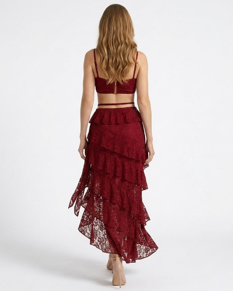 A woman shown from the back wearing a matching burgundy lace two-piece set with a ruffled tiered maxi skirt and a cropped bralette-style top, photographed against a plain light gray studio background.