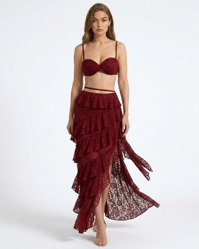 A woman modeling a deep burgundy lace two-piece set consisting of a bralette top and a tiered ruffled high-low skirt, photographed against a seamless white studio background.