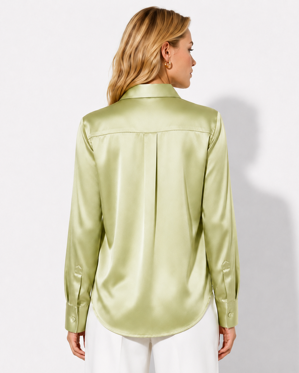 A woman shown from the back wearing a light olive green satin button-up blouse and white pants, photographed against a plain white studio background.