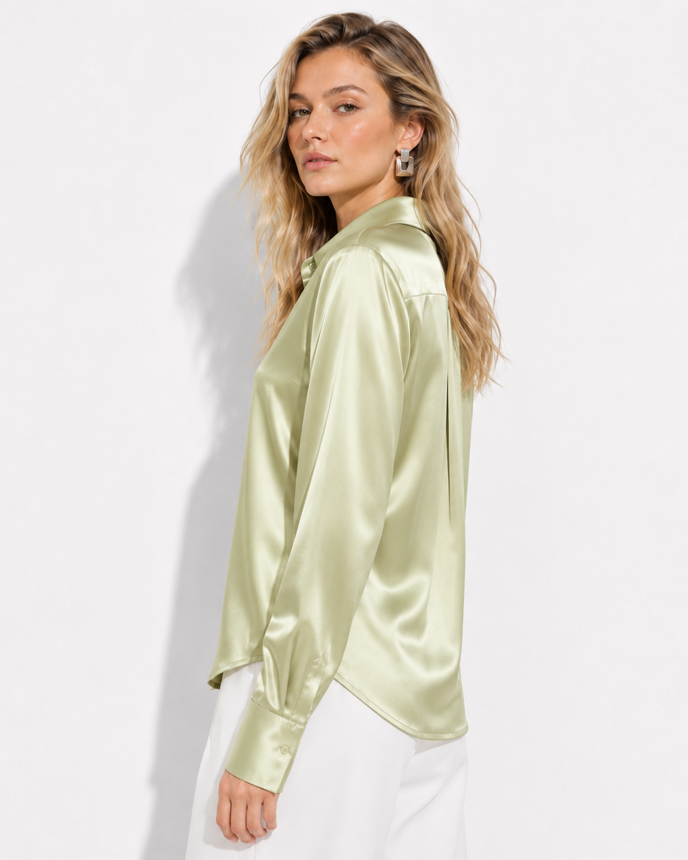 A woman wearing a shiny light sage green satin blouse with white pants, photographed against a clean white studio background.