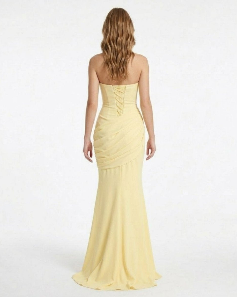 A woman viewed from the back wearing a strapless pale yellow mermaid-style gown with a lace-up corset back and a full floor-length skirt, photographed against a plain white studio background.