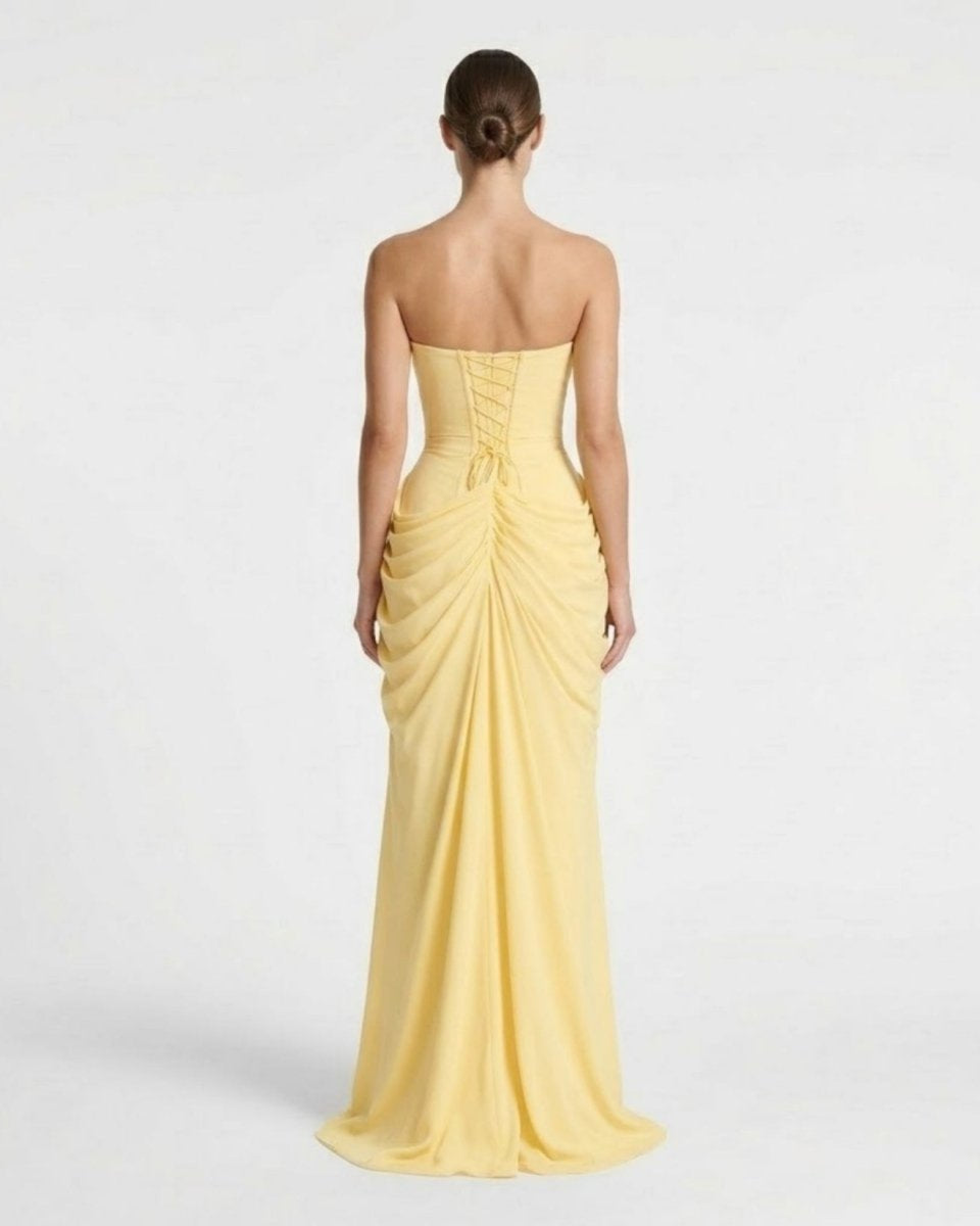 A woman viewed from the back wearing a strapless yellow floor-length gown with ruching and a gathered waist, photographed against a plain light gray studio background.