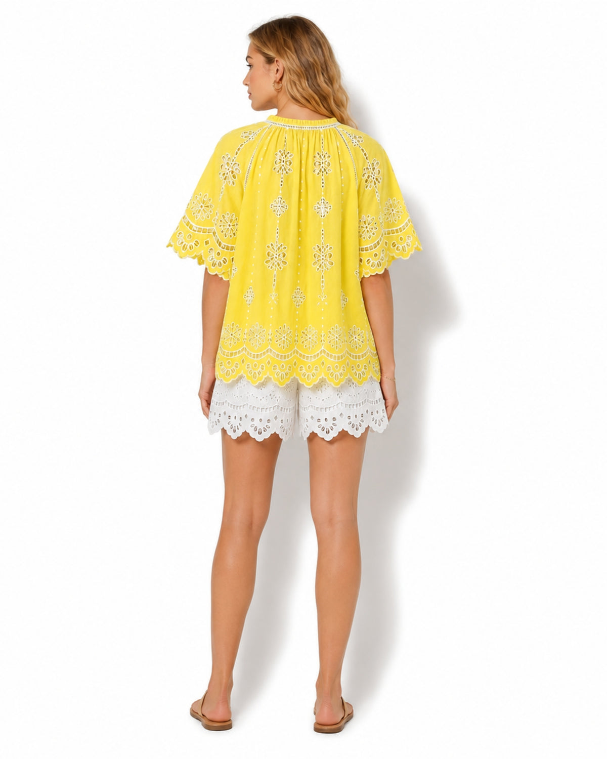 A woman shown from the back wearing a bright yellow embroidered short-sleeve top and white lace shorts against a plain white studio background.
