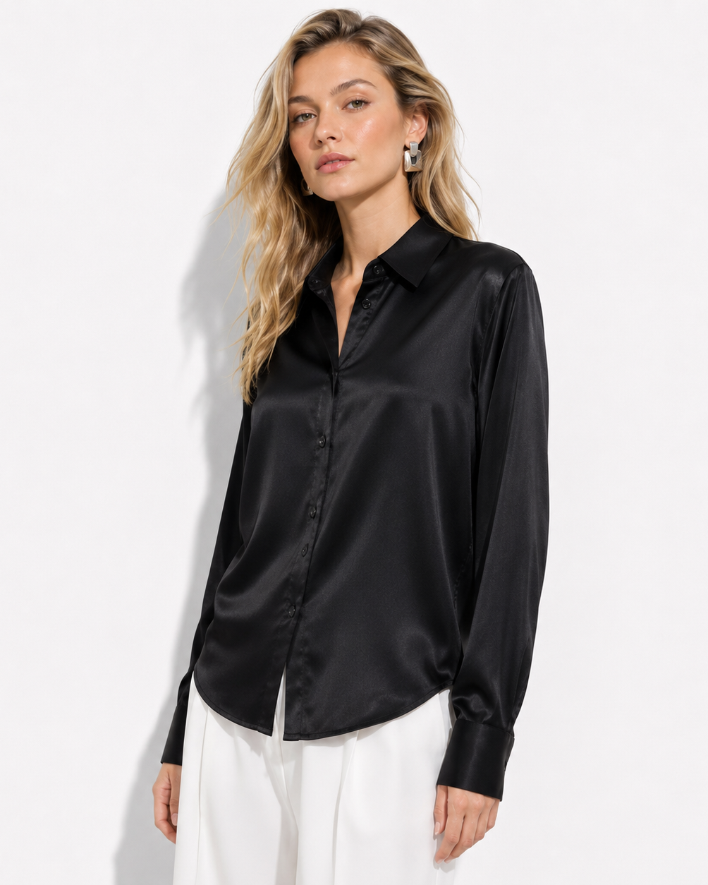 A woman modeling a black satin button-down shirt with white trousers, standing against a plain white studio background.