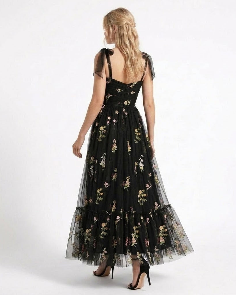 A woman modeling a long black tulle maxi dress with a floral print, shown from the back against a plain white studio background.