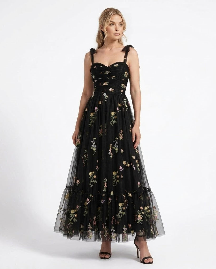 A woman modeling a black sleeveless maxi dress with a sweetheart neckline, floral print, and tiered tulle skirt against a plain white studio background.