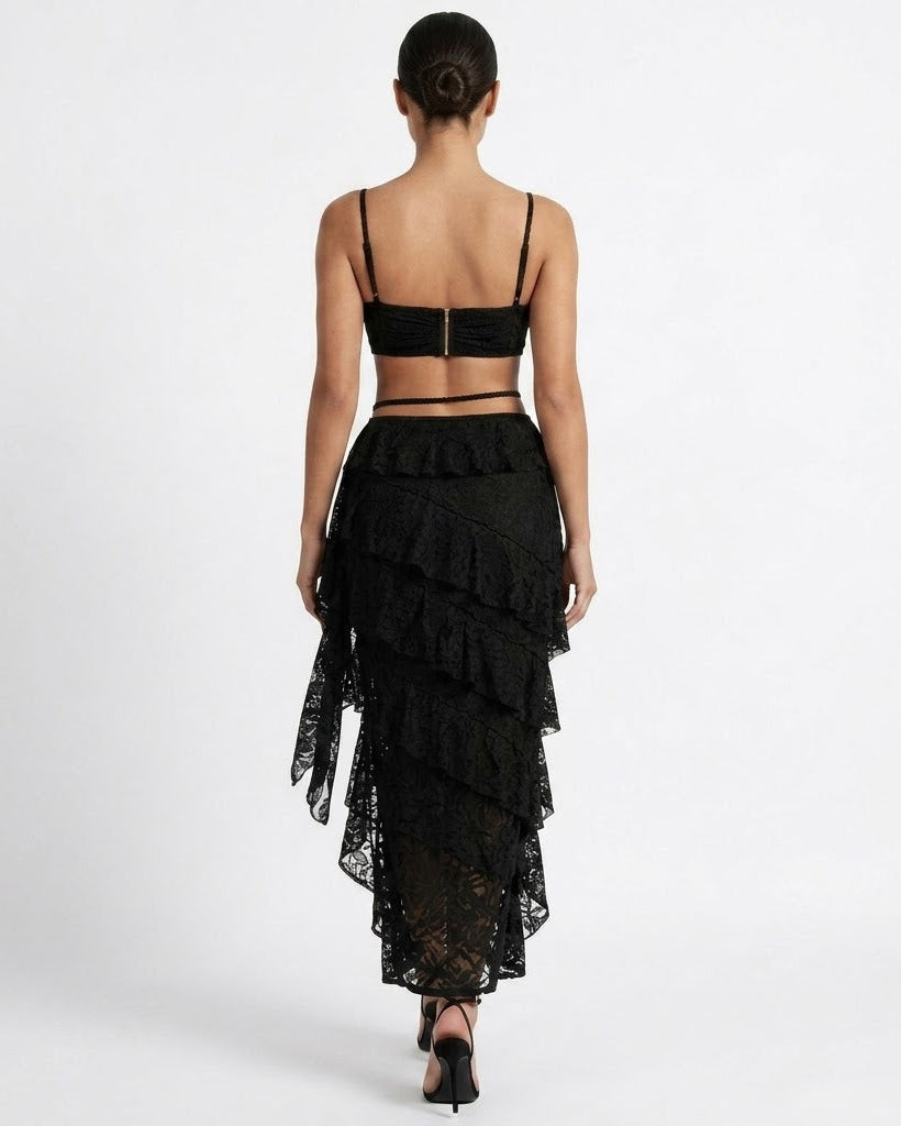 A woman shown from the back wearing a black strappy crop top and a black ruffled lace midi skirt with an asymmetrical hem, photographed against a plain light gray studio background.