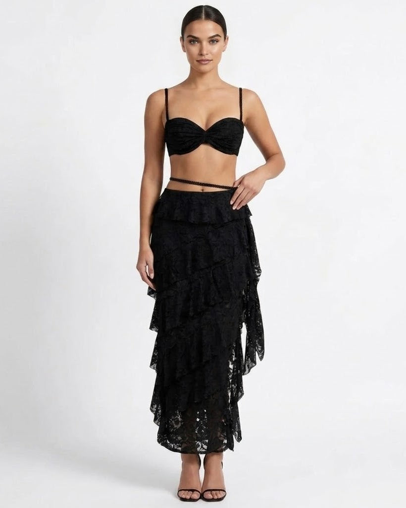 A model wearing a black lace two-piece outfit consisting of a bandeau-style crop top with thin straps and a long, tiered ruffled maxi skirt, paired with black strappy heels, against a plain white studio background.